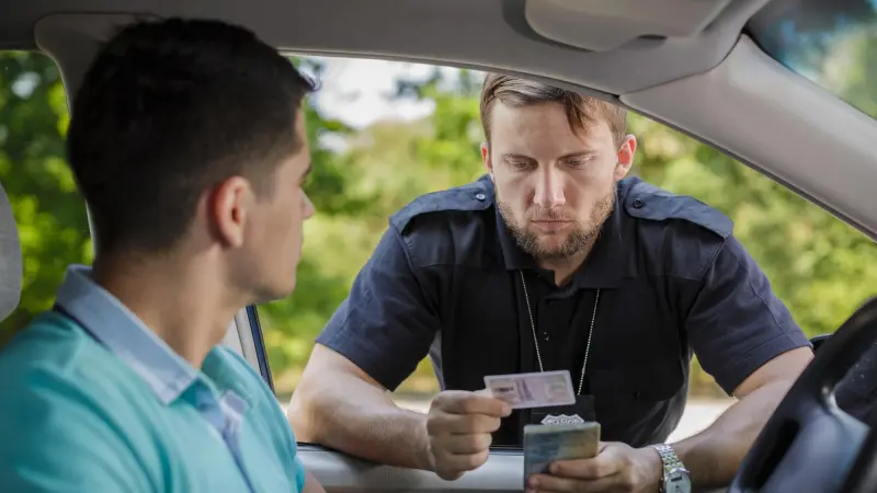 A police officer examines a driver's identification documents through the car window. The officer holds a driver's license and possibly a vehicle registration in hand, while attentively looking at the driver. This image highlights the need for rapid and reliable authentication of identity documents and driving permits to prevent fraud and counterfeiting, emphasizing the relevance of technologies like Origify for authorities and security services in ensuring document integrity.