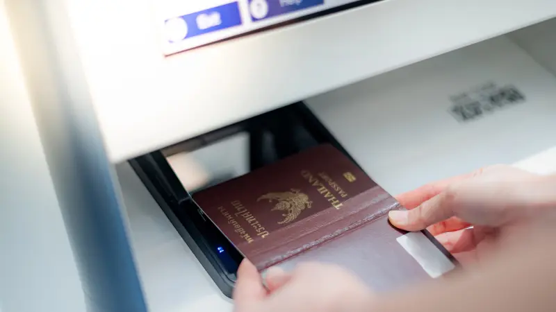 Close-up showing the process of scanning a Thai passport at an automated terminal or verification device. A hand places the passport onto a scanning surface, which is marked with indicators and numbered steps (1, 2, 3). This image illustrates the application of security technologies for fast and reliable authentication of travel documents at airports, border controls, or during identity verification, to detect counterfeits and improve process efficiency.