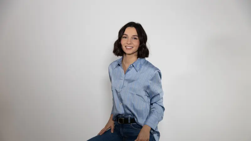 Portrait of a young, smiling woman with short, dark hair, dressed in a blue striped blouse and jeans, against a light gray background. The image conveys friendliness and competence, typical for a team member introduction, contact person, or brand ambassador of Origify, projecting the company's professionalism and approachability.