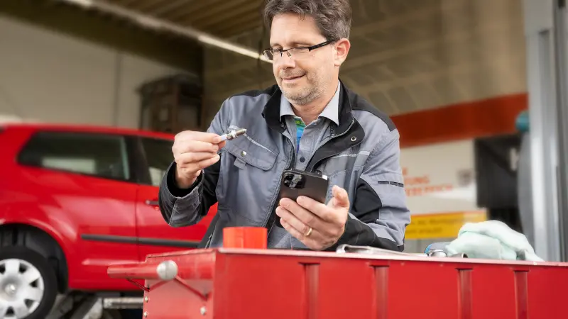 An automotive mechanic or technician in a workshop verifies the authenticity of a spark plug using a smartphone app. The image illustrates the application of Origify technology for authenticating spare parts, aiming to identify counterfeits and ensure quality and safety in vehicle maintenance. This emphasizes protection against fake auto parts and the safeguarding of product integrity.